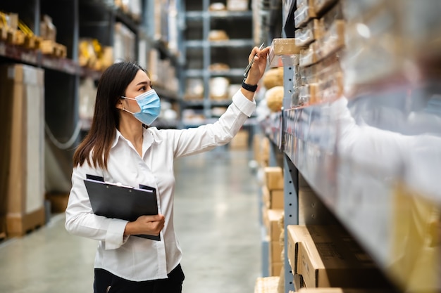 Woman worker with medical mask holding clipboard and checking inventory in the warehouse during coronavirus pandemic