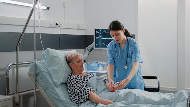 Woman working as nurse using oximeter for oxygen saturation measurement to cure elder patient in bed. Medical assistant putting tool on hand of woman with oxygen tube and IV drip bag