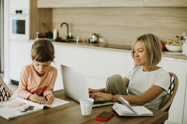 Woman working at home with her little daughter sitting near and drawing