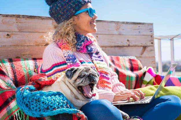 Woman working on laptop computer outdoor sitting on a wooden bench with her best friend old pug dog. Happy people with animals in winter season. Cheerful female and puppy friendship concept