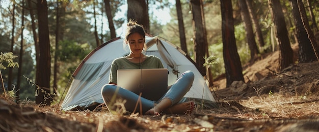 Woman working on a laptop in a forest sitting on the ground near a tent