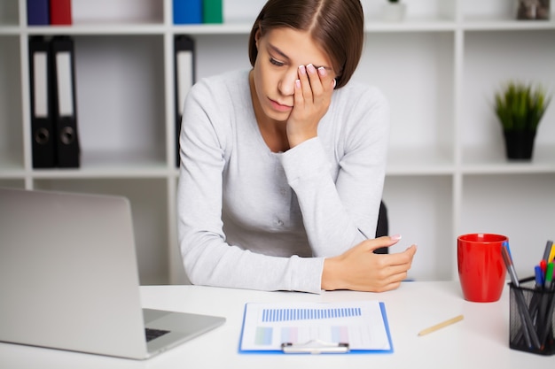 Woman working at office desk in front of laptop suffering from chronic daily headaches.