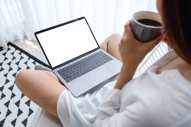  of a woman working and typing on laptop computer with blank screen while sitting and drinking coffee in a bedroom at home