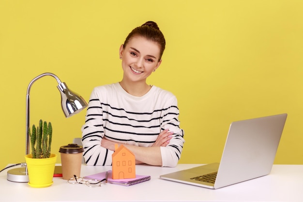 Woman at workplace with paper house looking at camera with smile dreaming about her own apartment