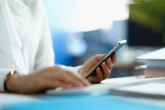 A woman works at a computer and holds a smartphone