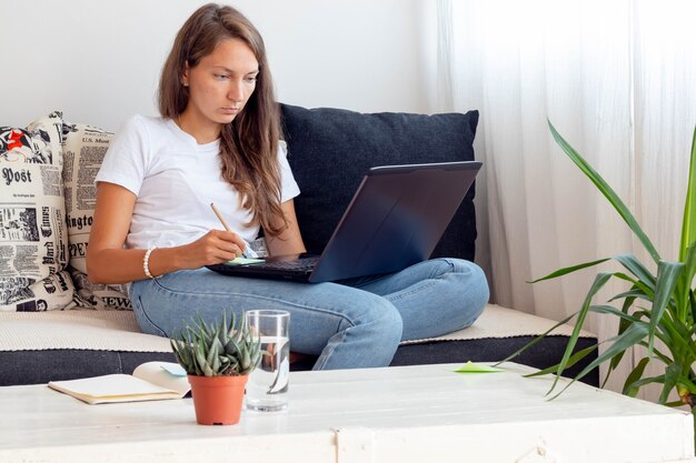 Woman works at home, seats close to Laptop with notebook, glass of water on table in home workspace