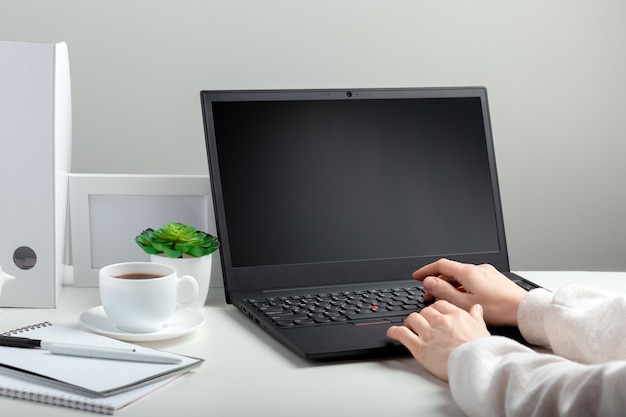 Woman works on laptop in online education on workspace. Female hands typing on laptop. Black laptop display blank. Workplace in home office for remote work minimal style on white wall.