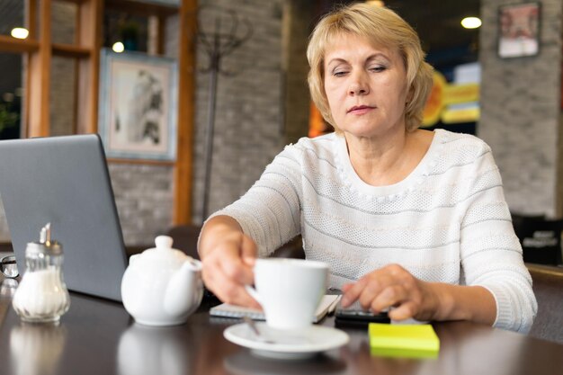 A woman works with a laptop at a table in a cafe or in the office. A middle-aged woman works remotely. She's a businesswoman.