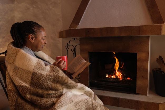 Woman wrapped in a blanket drinking a hot chocolate by the fireplace in winter Concept indoors senior life relaxation