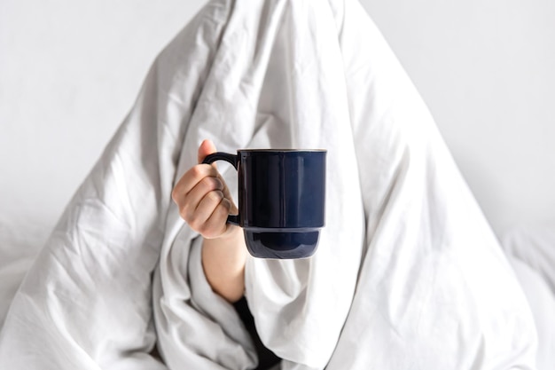 A woman wrapped in a blanket sits in bed with a cup of coffee in her hands