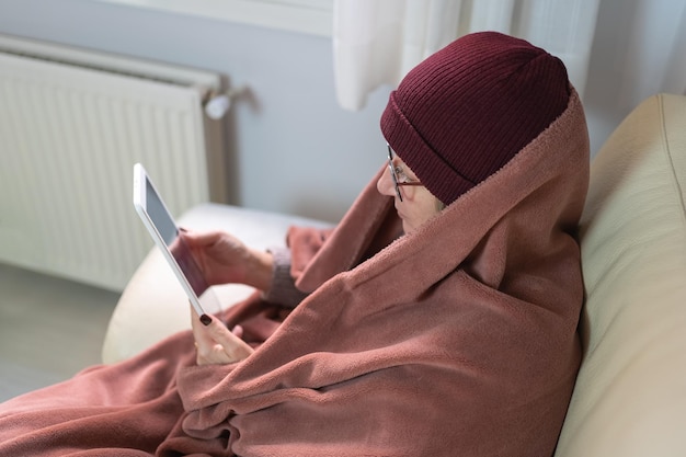 Woman wrapped in a blanket and wearing a wool hat looking at a tablet while warming up by the cold of winter and energy savings