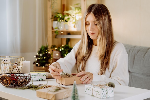 Woman wrapping christmas eco natural gift boxes at home preparing presents on white table with decor