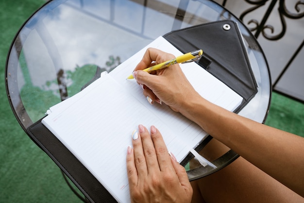 Woman writes in a notebook with a pen while sitting at the table at home on the terrace