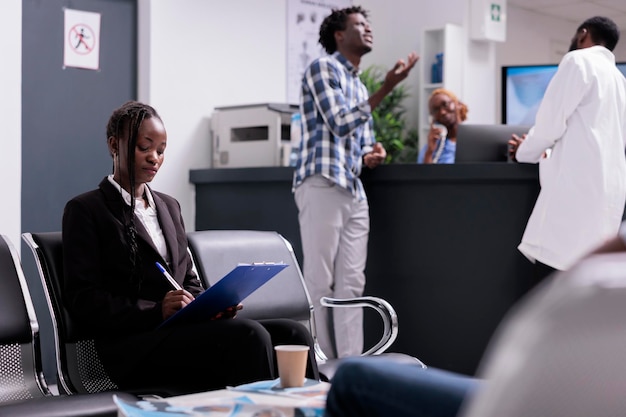 Woman writing medical report papers and checkup form to receive healthcare insurance support before appointment with physician. Patient sitting in waiting room area at hospital facility lobby.