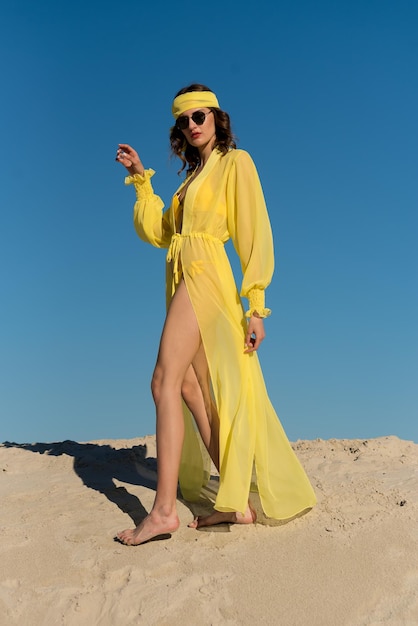 A woman in a yellow dress stands in the sand and wears a long yellow dress.