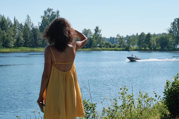 Woman in yellow dress stands on the shore and looks at floating boat