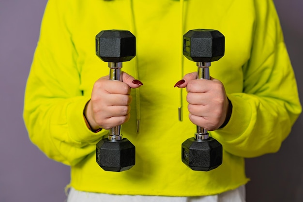 Photo woman in yellow hoodie lifting dumbbell against a purple wall