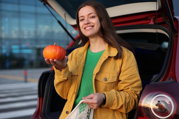 Woman in yellow jacket hold orange pumpkin and shopping bag with a groceries sitting on red cars tru