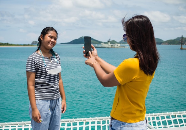 A woman in a yellow shirt is holding a phone to take pictures of a girl in a gray shirt With a background of blue water and small mountains It is tourism after the corona virus outbreak