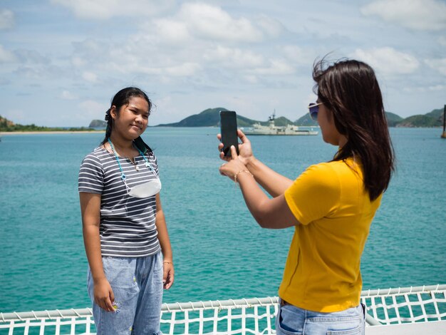 A woman in a yellow shirt is holding a phone to take pictures of a girl in a gray shirt With a background of blue water and small mountains It is tourism after the corona virus outbreak