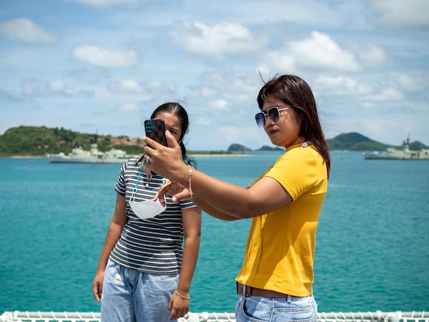 A woman in a yellow shirt is holding a phone to take a selfie with a girl in a gray shirt with a background of blue water and small mountains It is tourism after the corona virus outbreak