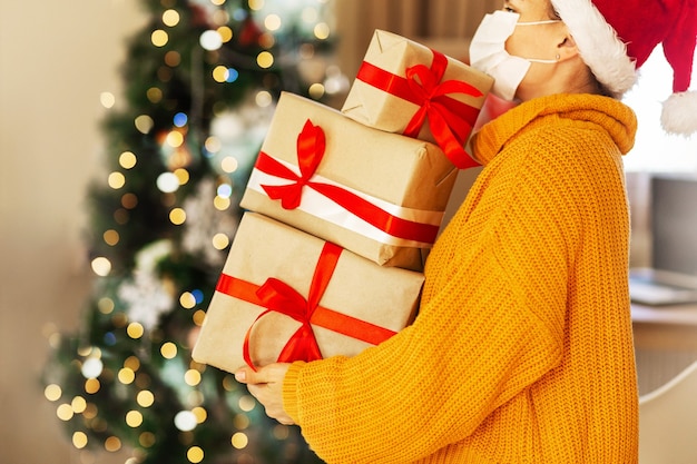 Woman in yellow sweater in protective medical mask and santa claus hat holding boxes delivery