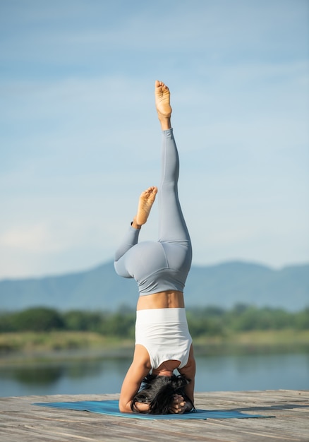 Woman on a yoga mat to relax in the park. Young sporty asian woman practicing yoga, doing headstand exercise, working out, wearing sportswear, pants and top.