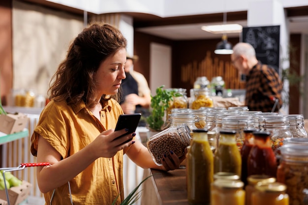 Woman in zero waste store using smartphone notes app to check shopping list Meticulous customer looking to replenish pantry at home with organic chemicals free food essentials