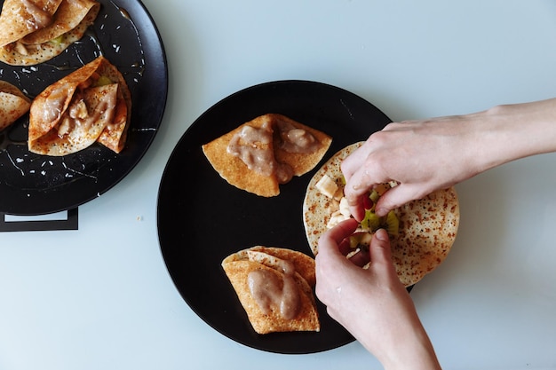 Womans hand decorates pancakes with a fruits kiwi and banana Black plate white table