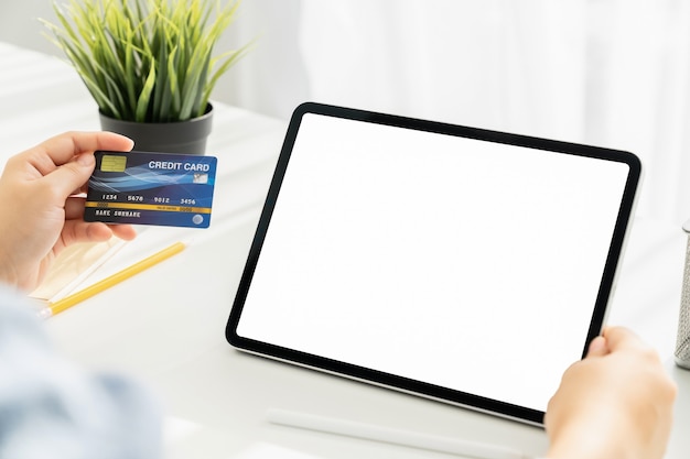 Womans hand holding a credit card and using a tablet for online shopping on a white desk