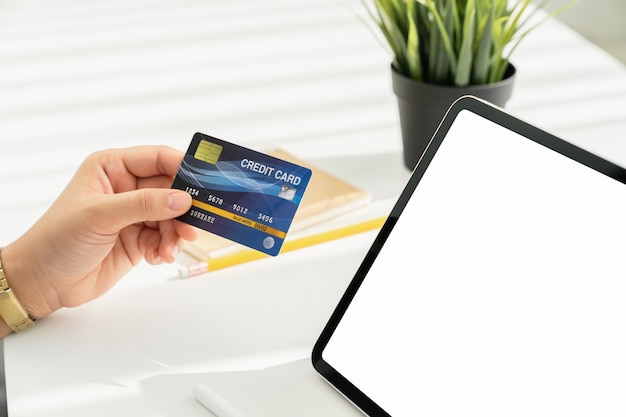 Womans hand holding a credit card and using a tablet for online shopping on a white desk