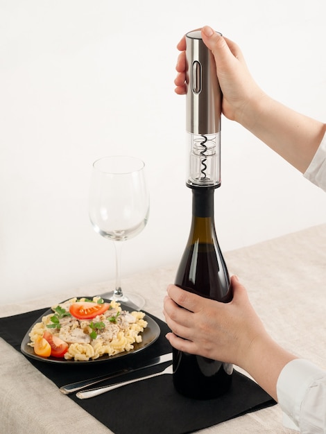 A womans hand opens a bottle of wine with an electric corkscrew made of gray metal