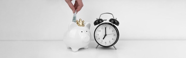 Womans hand put dollar bancnote into a piggy bank with alarm clock