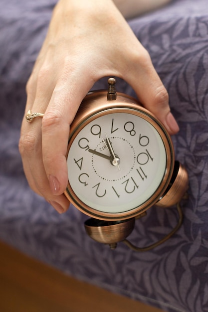 A womans hand with an alarm clock hanging from the bed The concept of a difficult awakening an early rise