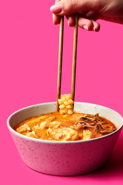 A womans hand with chopsticks takes seafood from the ramen Pink background