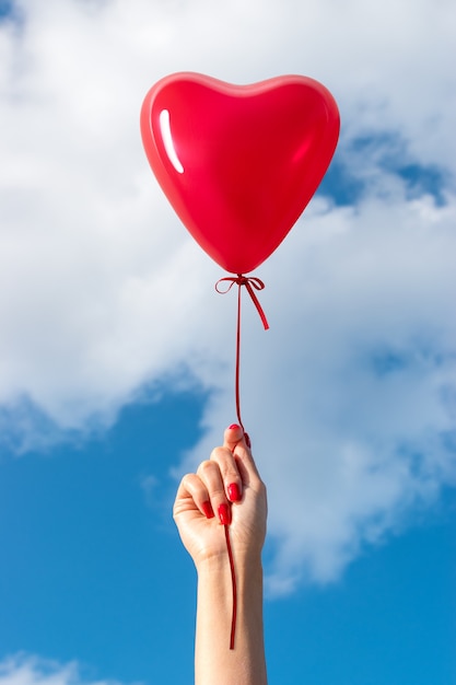 Womans hand with heart shaped balloon on background of sky.