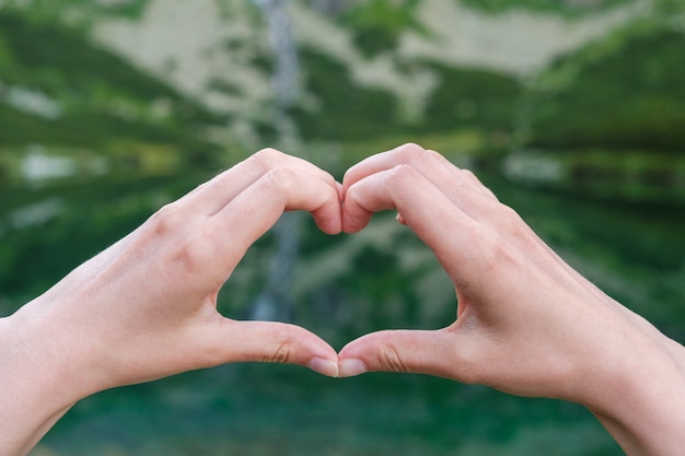 Womans making heart shape with her hands against the mountains near to the lake.