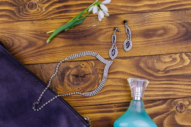 Women accessories on wooden background Clutch bag bottle of perfume necklace earrings and snowdrops on wood table Beauty and fashion composition Top view flat lay