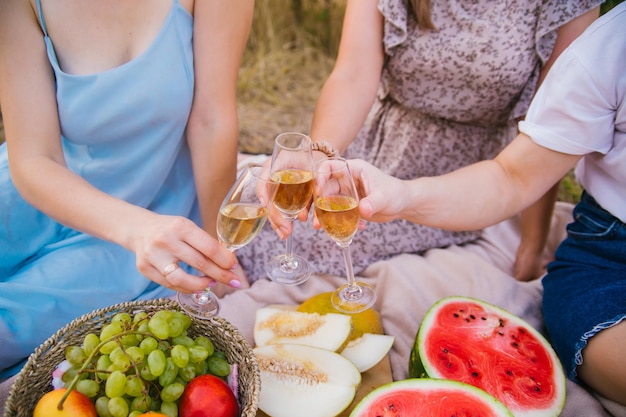 Women clink glasses with white wine or champagne at an outdoor party Women's hands hold transparent glasses with an alcoholic drink at a picnicCloseup Juicy fruits in the background