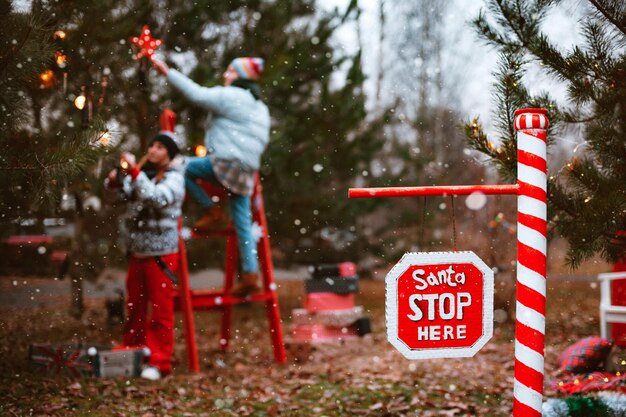 Women decorate a christmas tree outdoors sparkler lights up a stop sign with the text Santa stop here.