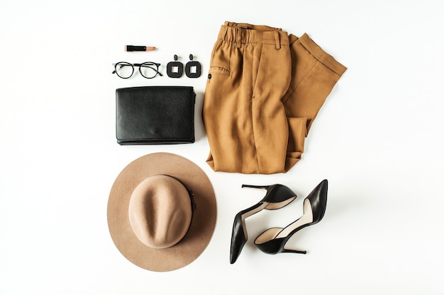 Women fashion clothes, accessories on white. Flat lay, top view minimal trendy lifestyle collage. Pants, high-heel shoes, glasses, hat, purse bag, earrings