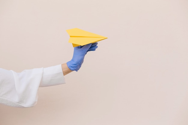 Women hands in a protective blue latex gloves holding yellow paper airplane on beige background. Copy space