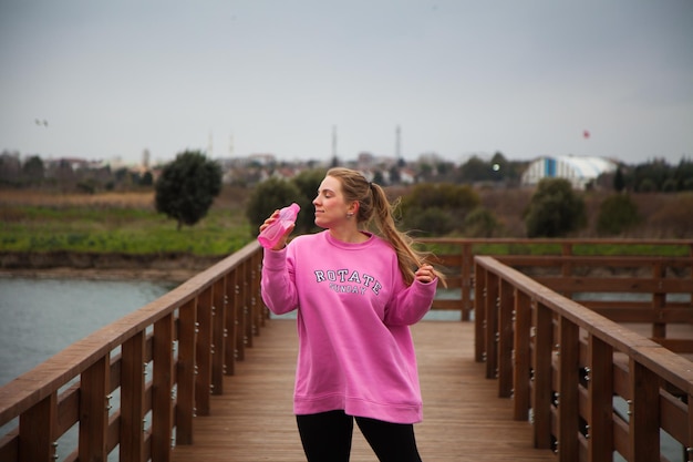 Women in pink hoodie doing exercises outdoors in the pier in autumn . Woman drinking water after exe