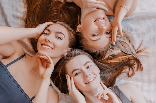 Women rest and have fun. Girlfriends laugh at home lying on the floor on pillows. Tree girls make homemade face and hair beauty masks. Women take care of youthful skin.