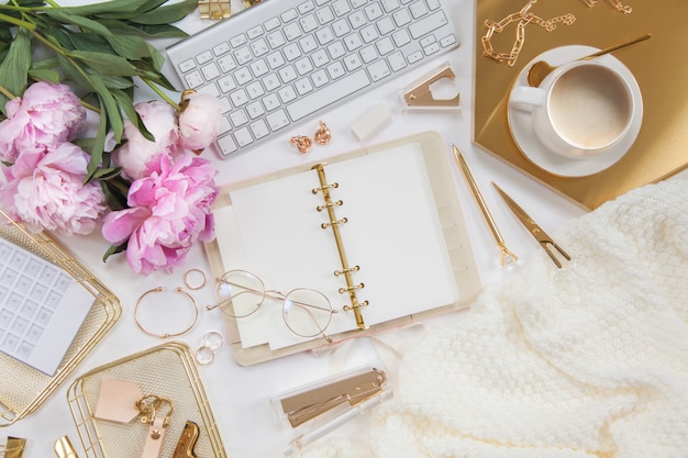 Women's diary and golden stationery. Bouquet of pink peonies. Glasses, a white keyboard, pen, scissors and coffee on the desktop.