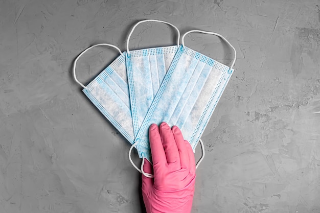 Women's hand in pink gloves hold three blue medical disposable mask on grey background. Hands of woman with mask close-up. Top view, flat lay.