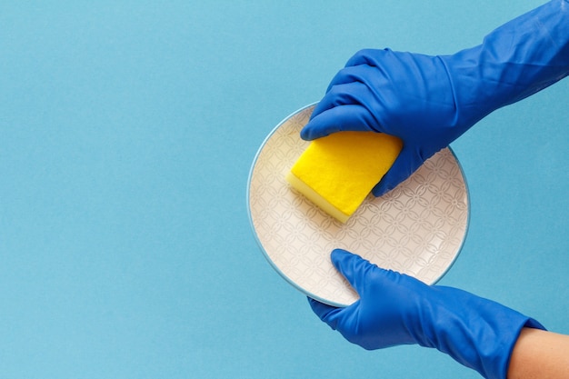 Women's hands in blue protective glove with plate and sponge on blue background. Washing and cleaning concept.