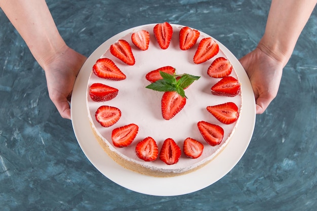 Women's hands hold delicious cheesecake with fresh strawberries and mint homemade recipe without baking on a dark blue stone table Copy space