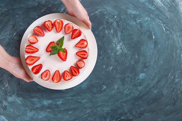 Women's hands hold delicious cheesecake with fresh strawberries and mint homemade recipe without baking on a dark blue stone table Copy space