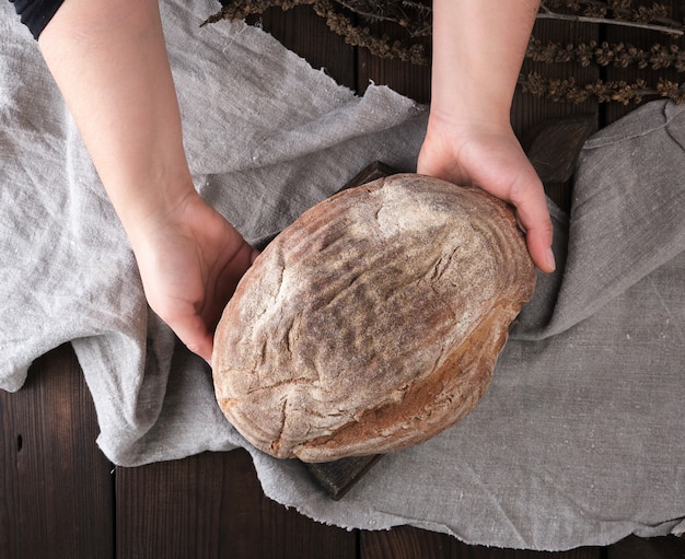 Women's hands hold a whole round loaf of rye flour bread 